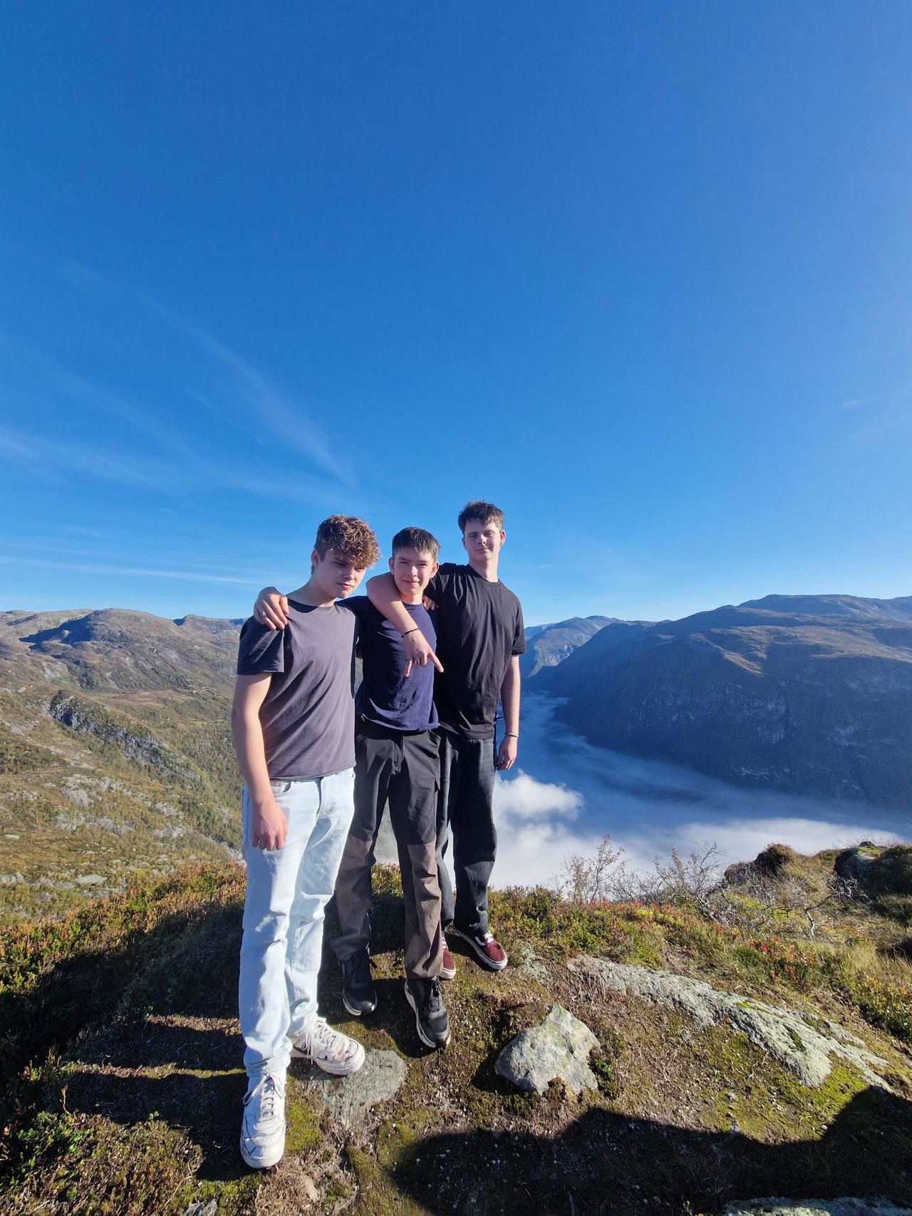 Tre gutar står på toppen av eit fjell med blå himmel i bakgrunn