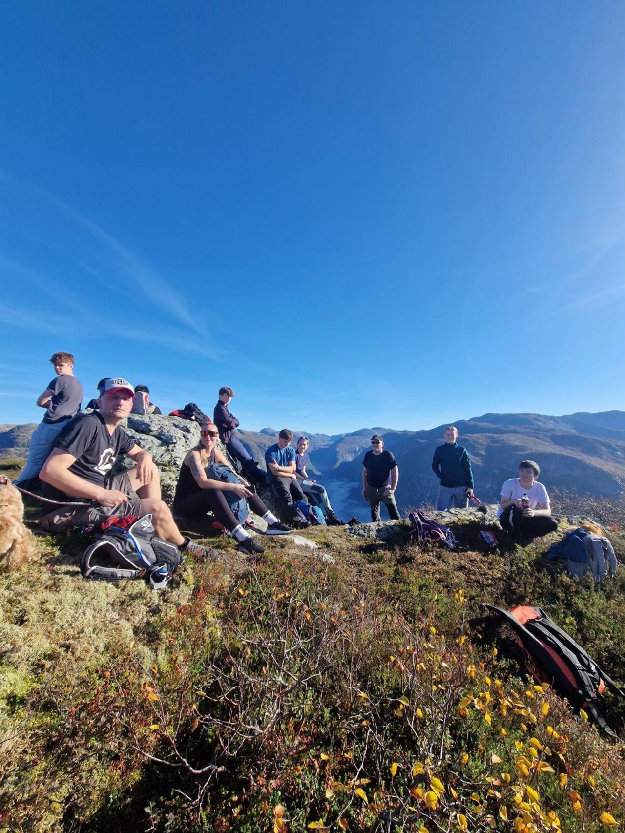 Fleire elevar sit og har pause på toppen av eit fjell med blå himmel i bakgrunn