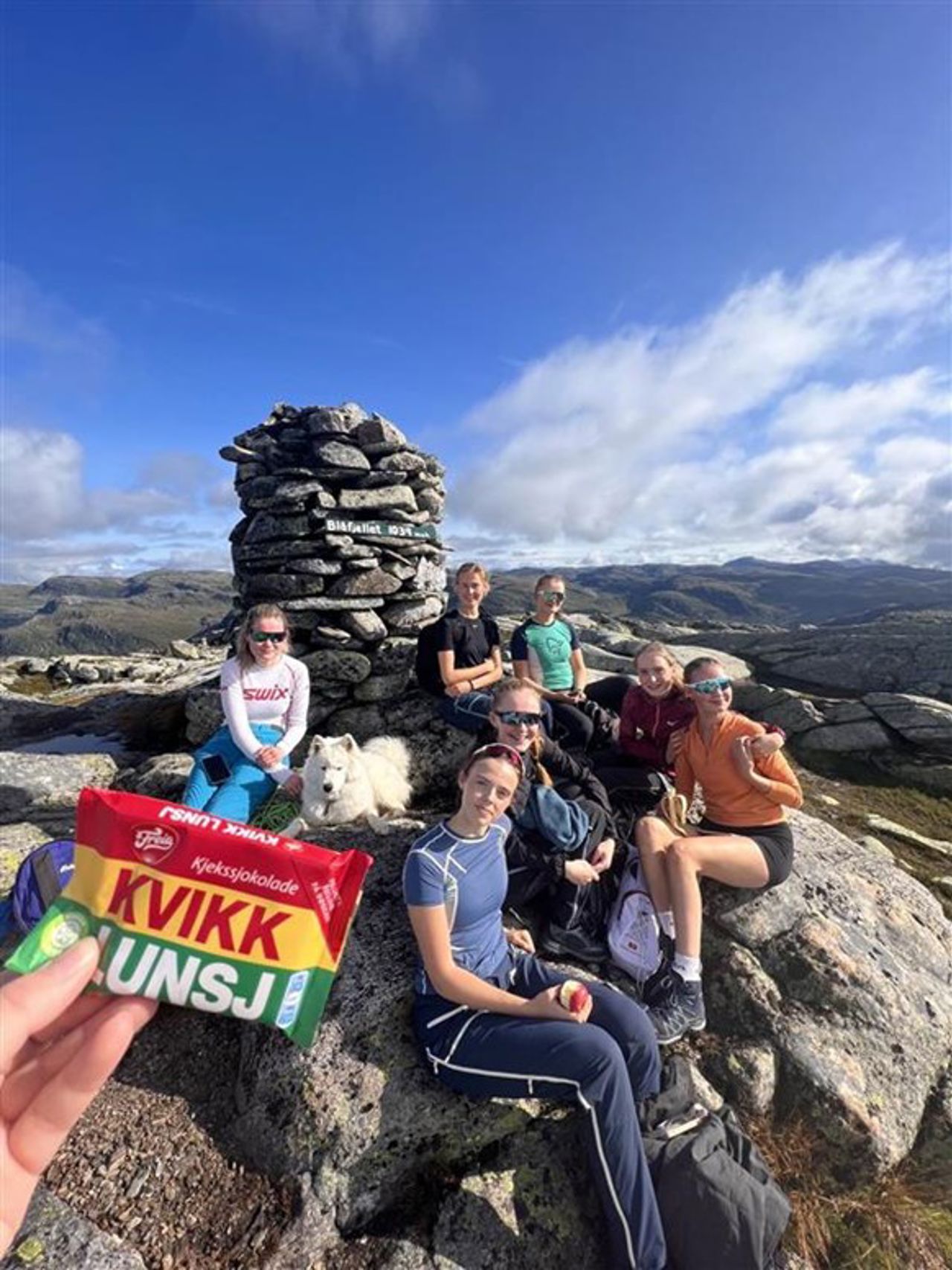 Ei elev held ein pakke med kvikk lunsj i handa foran ein varde på fjellet. Sju jenter sit framfor varden.
