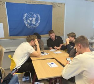 Fire elevar sit rundt eit bord og diskuterer. FN flagget heng i bakgrunn