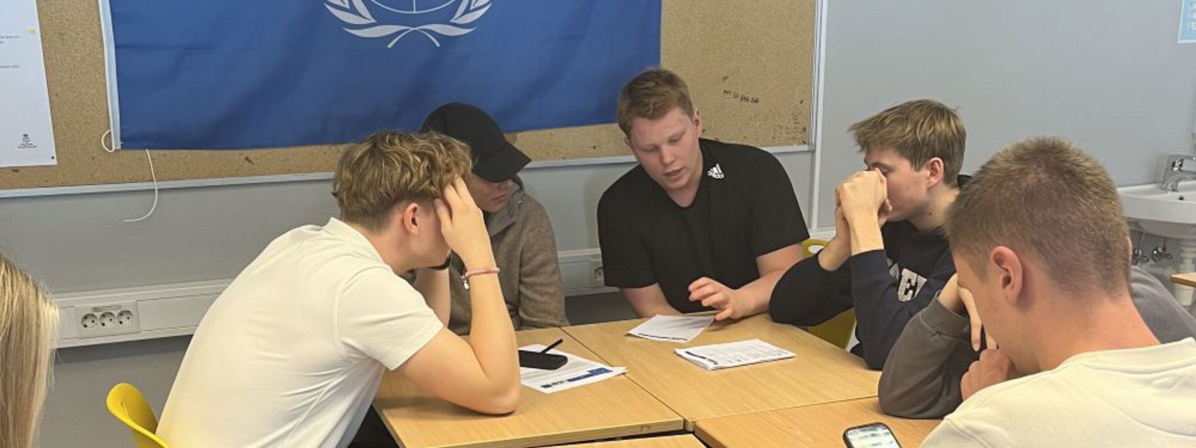 Fire elevar sit rundt eit bord og diskuterer. FN flagget heng i bakgrunn